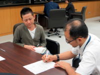 医師と患者が対話しながら書類に記入している様子。背景には他の人々がいる。