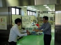 医療施設内で、白衣を着た二人の男性が何かを手渡ししている様子。背景には医療機器が見える。