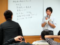 講義中の男性がホワイトボードを指し示し、参加者が聴いている様子。