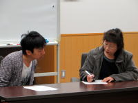 学生と教員が会話を交わしながら、メモを取る様子が写った会議室の風景。