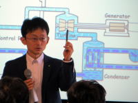 講演中の男性が手にペンを持ち、背景には発電システムの図が描かれたスライドが映し出されています。
