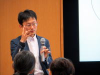 福島県立医科大学での講演中、マイクを持って話す男性の姿。
