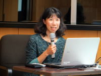 講演を行う女性がマイクを持ち、パソコンの前で話している様子。背景には会議室の一部が見える。