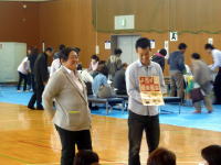 福島県立医科大学のイベントで、参加者に資料を配布する男性とそれを見守る女性。背景には多くの人々が集まっている。