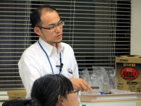 講義中の男性が聴衆に向かって話している様子。背景には窓と資料が置かれた机が見える。