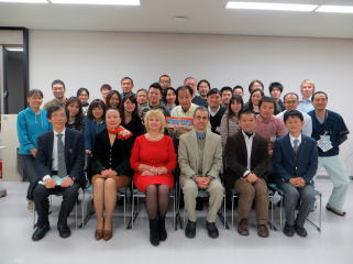修了式の参加者たちが集合し、笑顔でポーズをとっている様子。前列には数名の人物が座り、後列には多くの人々が立っている。