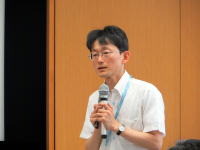 講演中の男性がマイクを持ち、聴衆に向かって話している様子。背景には木目調の壁が見える。