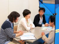 医療に関する話し合いを行う4人の男女がテーブルを囲んでいる様子。背景には青いテントが見える。