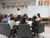 大学の教室でプレゼンテーションを行う女性と、聴講する学生たちの様子。背景には歴代の教授の肖像画が飾られている。