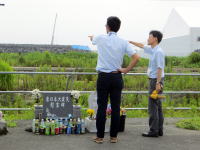 福島県立医科大学の前で、二人の男性が指を指しながら話している様子。周囲には花や飲料が供えられている。