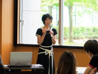 講演を行う女性がマイクを持ち、聴衆に向かって話している様子。背景には緑の木々が見える。
