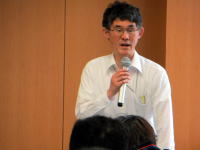 講演を行う男性がマイクを持ち、聴衆に向かって話している様子。背景には木目調の壁が見える。