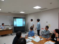 会議室でプレゼンテーションを行う学生たちと、資料を見守る教員の様子。