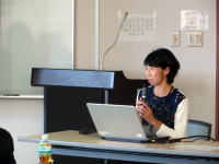 講演を行う女性がマイクを持ち、パソコンの前で話している様子。背景にはホワイトボードが見える。