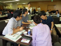 大学の教室でグループディスカッションを行う学生たちの様子。テーブルには資料や飲み物が置かれている。