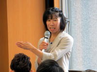 講演中の女性がマイクを持ち、参加者に向かって話している様子。背景には窓があり、明るい室内の雰囲気。