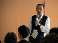 講演を行う男性がマイクを持ち、聴衆に向かって話している様子。背景には木製の壁が見える。