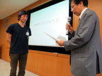 福島県立医科大学での表彰式の様子。スーツを着た男性がマイクを持ち、受賞者に向かって話している。