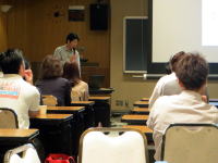 講義を受ける学生たちとプレゼンテーションを行う講師が写る教室の風景。