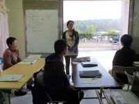 教室で講義を受ける学生たちと、前で話す女性講師の様子。窓の外には緑豊かな風景が広がっている。