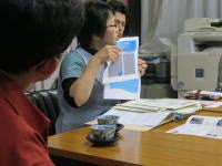 会議中の女性が資料を指し示し、他の参加者がそれを見つめている様子。テーブルには書類や飲み物が置かれている。