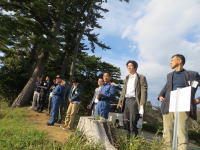 福島県立医科大学の関係者が屋外で集まり、何かを指示している様子。背景には青空と木々が広がっている。
