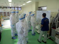 医療現場で防護服を着たスタッフがホワイトボードを囲んで説明を受けている様子。