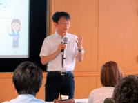 福島県立医科大学で講演を行う男性がマイクを持ち、聴衆に向けて話している様子。