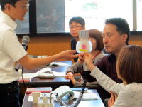医療に関する講義中、講師が参加者に資料を示している様子。背景には他の参加者が座っている。