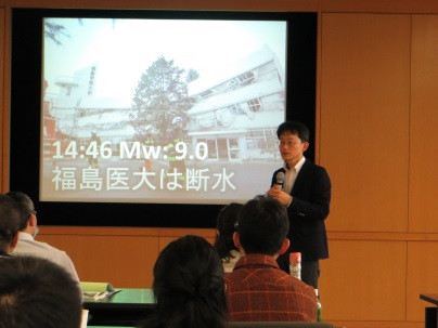 福島県立医科大学での講演中の男性と、震災に関するスライドが映し出された様子。