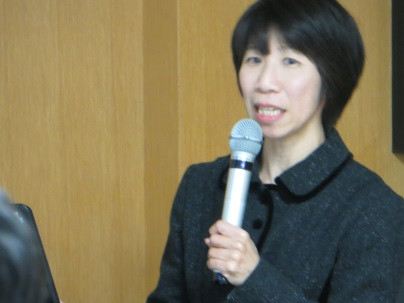 講演中の女性がマイクを持ち、聴衆に向かって話している様子。背景は木製の壁。