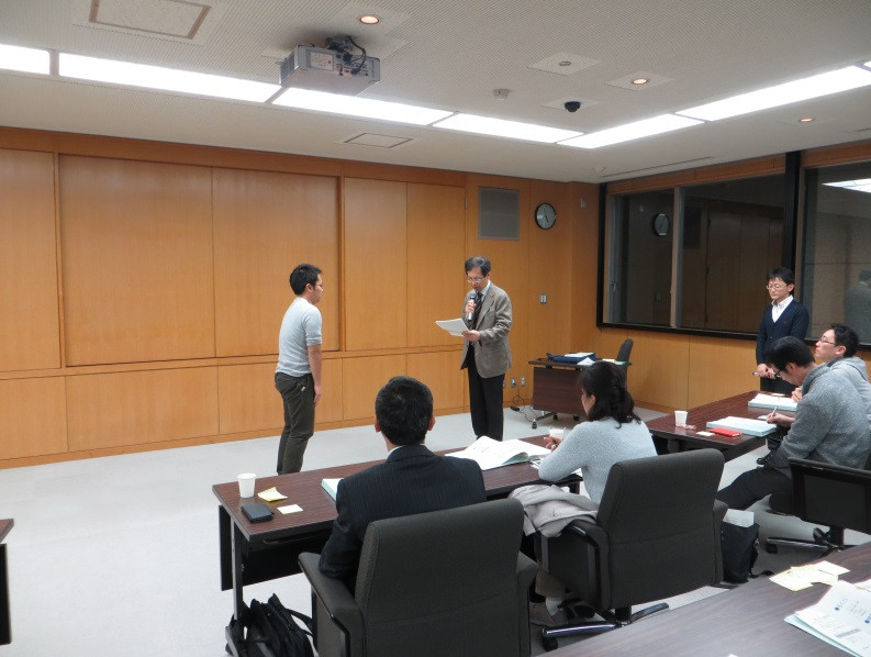 福島県立医科大学の教室で、講師が学生に指導している様子。学生は立っており、他の学生たちが座って見守っている。