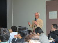 講演を行う白髪の男性が、聴衆に向かって話している様子。背景には参加者が座っている。