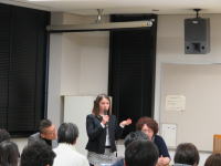 医療に関する講演を行う女性が、聴衆に向かって話している様子。背景には黒いカーテンとスピーカーが見える。