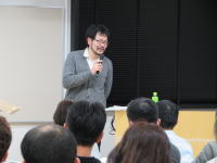 講演を行う男性がマイクを持ち、聴衆に向かって話している様子。背景には黒いカーテンがあり、参加者が数名見える。