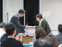 福島県立医科大学の授賞式で、男性が女性に賞状を手渡している様子。観衆が拍手を送る中、感動的な瞬間が捉えられている。