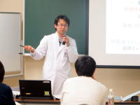 医療に関する講義を行う白衣を着た講師と、聴講する学生たちの様子。