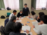 会議室でテーブルを囲んで話し合う8人の参加者。中央に立つ男性が発表している様子。