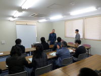 福島県立医科大学の教室で、講師が参加者に向けて話している様子。参加者はテーブルに座り、講義を聴いている。