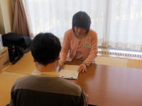 医療相談を行う女性とその向かいに座る男性の後ろ姿。明るい窓辺での対話の様子。