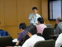 講義中の講師が参加者に向けて話している様子。背景には木目調の壁と窓が見える。