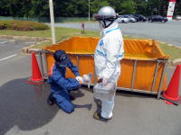 防護服を着た作業員が、別の作業員に器具を手渡している様子。背景にはオレンジ色のバリアとコーンが見える。