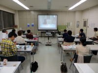 教室でプレゼンテーションを受ける学生たちの様子。スクリーンには映像が映し出されている。
