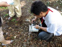 地面に座り込んで機器を操作する男性と、その周囲の人々が写っている風景。