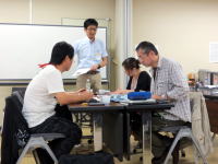 大学の教室で、数人の学生がテーブルを囲んで学習している様子。講師が立って説明している。