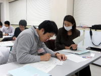 学生が教室で資料に取り組んでいる様子。手前の男性は集中して書き込みをし、女性は資料を見つめている。