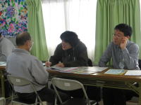 医療に関する相談を行う場面。テーブルを囲む3人の男性が話し合っている。背景にはカーテンと色とりどりのアートが見える。