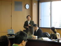 福島県立医科大学の会議室で講演を行う男性と参加者たちの様子。