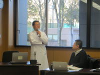 医師が講演を行い、聴衆が話を聞いている会議室の様子。
