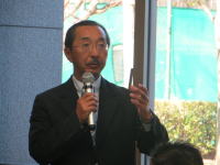 講演を行う男性がマイクを持ち、聴衆に向かって話している様子。背景には窓と緑の景色が見える。